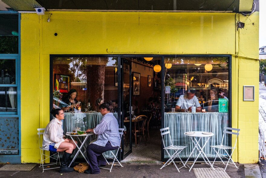 The exterior of a small wine bar painted yellow. There is a couple seated at the front drinking wine. There are people indoors seated at the window.