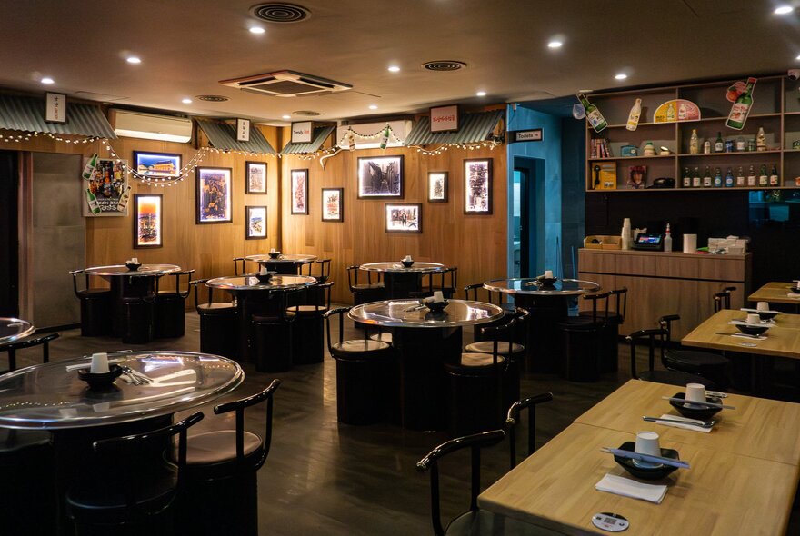 The interior of a restaurant with tables and chairs arranged in the space, small pictures hung on the wood panelled walls, and downlights.