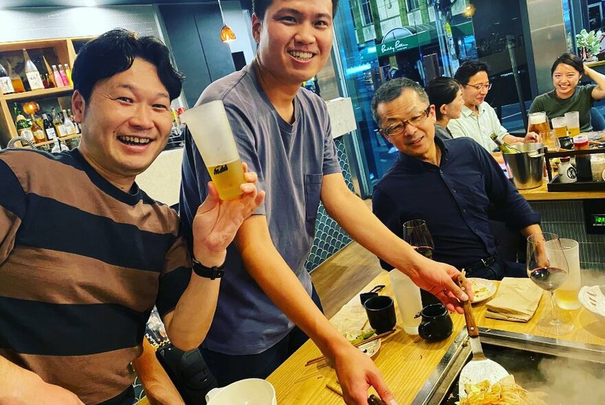 Dining patrons smiling and watching someone cook food on a hotplate in the centre of a table, inside a restaurant.