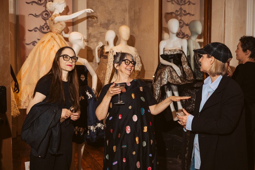 Three glamorous fashion personalities enjoying a drink with stylishly dressed mannequins on display behind them.