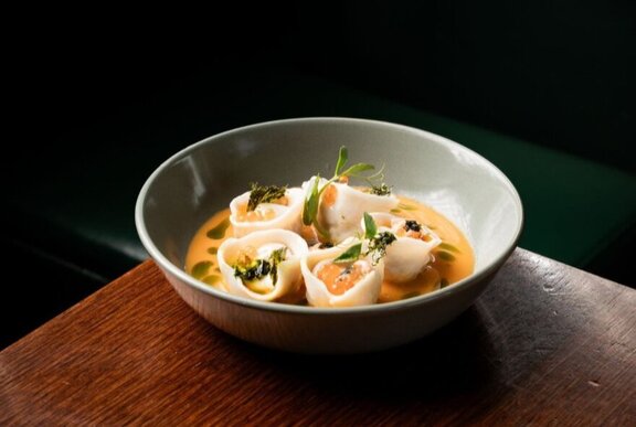 A bowl of noodles and eggs in a broth, resting on a wooden table.