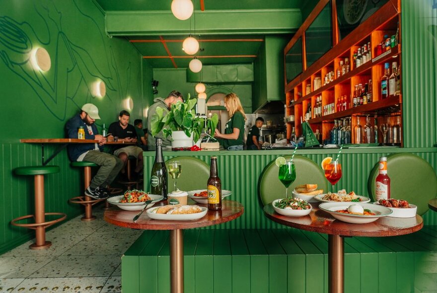 Interior of a small restaurant with high tables and stools against one wall, green walls, seats, and decor, people working behind a service area, and dishes of food and drinks arranged on two small tables in the foreground.