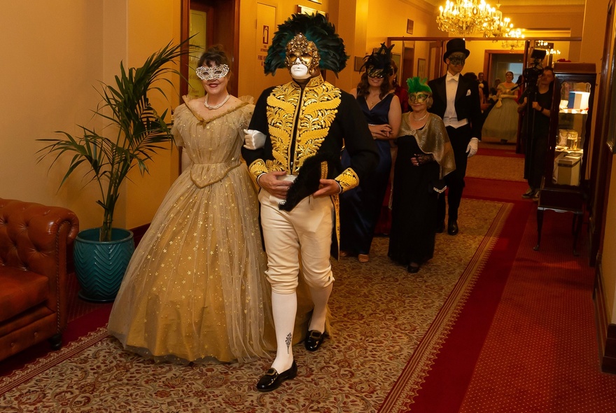 A couple wearing period costumes wearing face masks, walking down a carpeted hallway in a grand building.