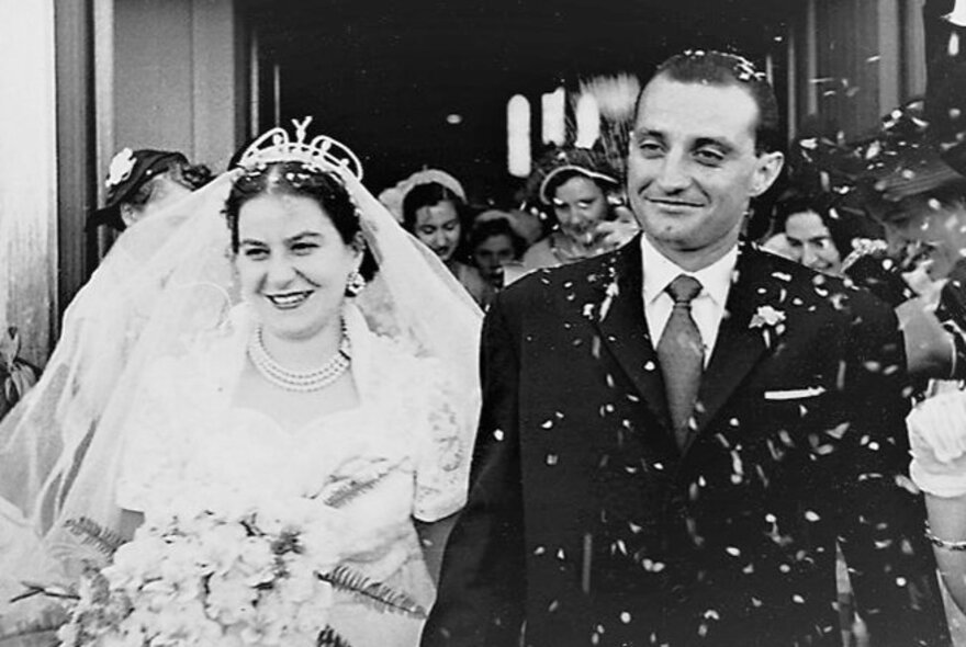 A vintage black and white photo dating from the mid-1950s, of a bride and groom leaving a church and being showered in confetti by onlookers.
