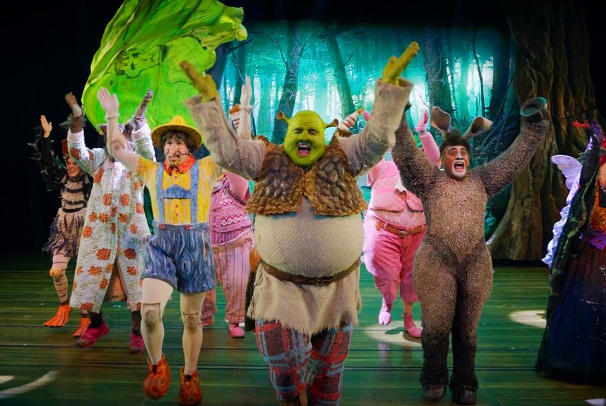 The cast of Shrek The Musical on a theatre stage dancing with their arms raised in the air.