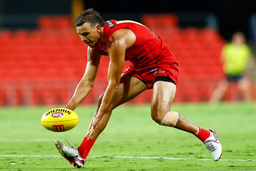 A Gold Coast AFL footballer leaning forward while running to grab a ball off the ground during a game.
