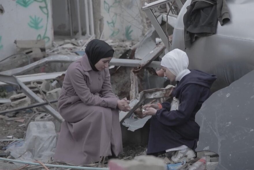 Women wearing hijabs weeping, seated on rocks in the ruins of a destroyed house.