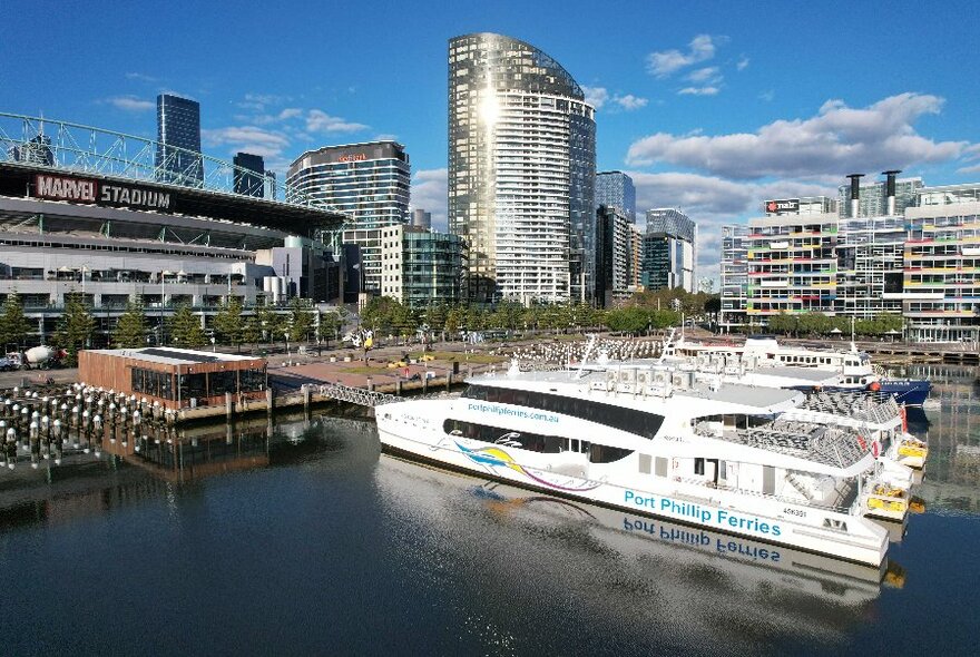 Aerial view of the Melbourne Ferry Terminal, with a large ferry docked at the harbour, and a view of Marvel Stadium and city buildings in the background.