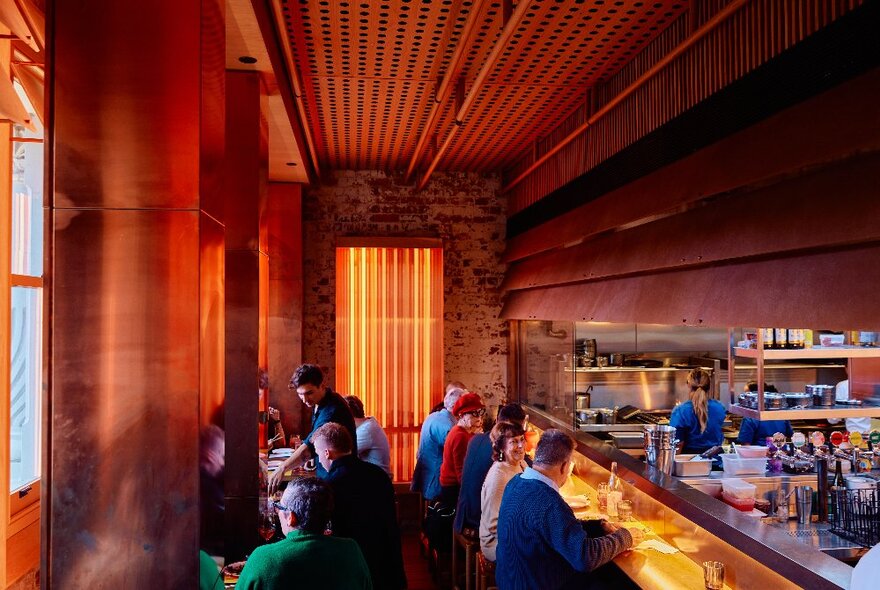 The interior of a restaurant with patrons seated at tables and at the bar in front of the kitchen.