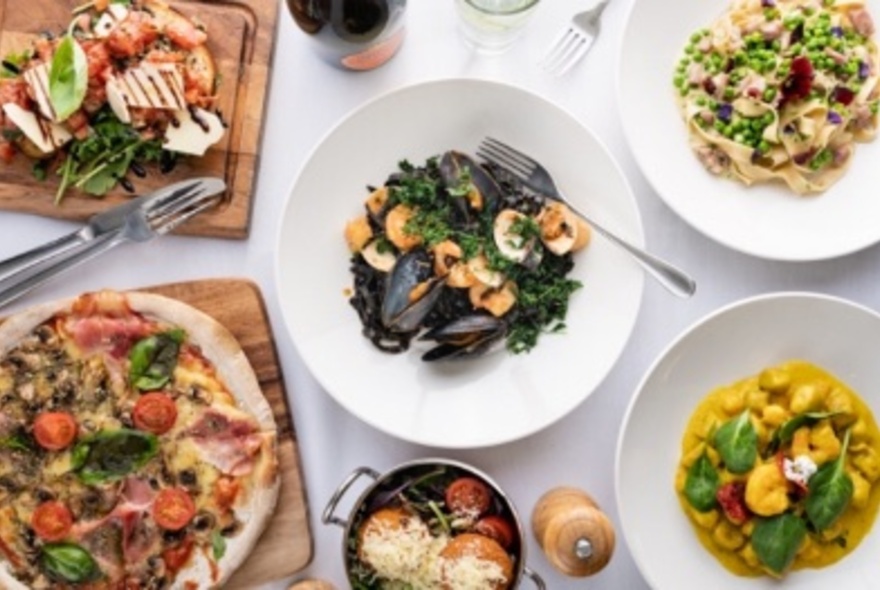 Selection of Italian dishes in white plates and wooden trays, on white table cloth, from above.