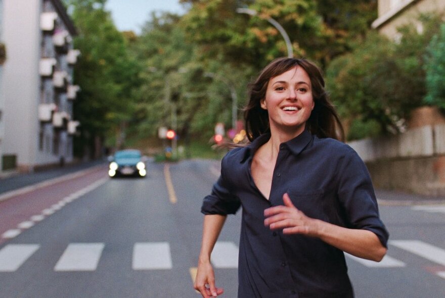 A scene from a film showing a woman smiling as she runs down the middle of a street, a zebra crossing and a car coming behind her. 
