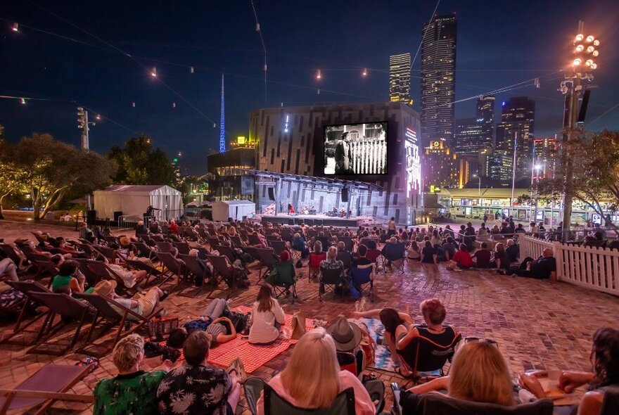 A seated audience in deck chairs at Fed Square at night, watching a silent movie accompanied by live music.