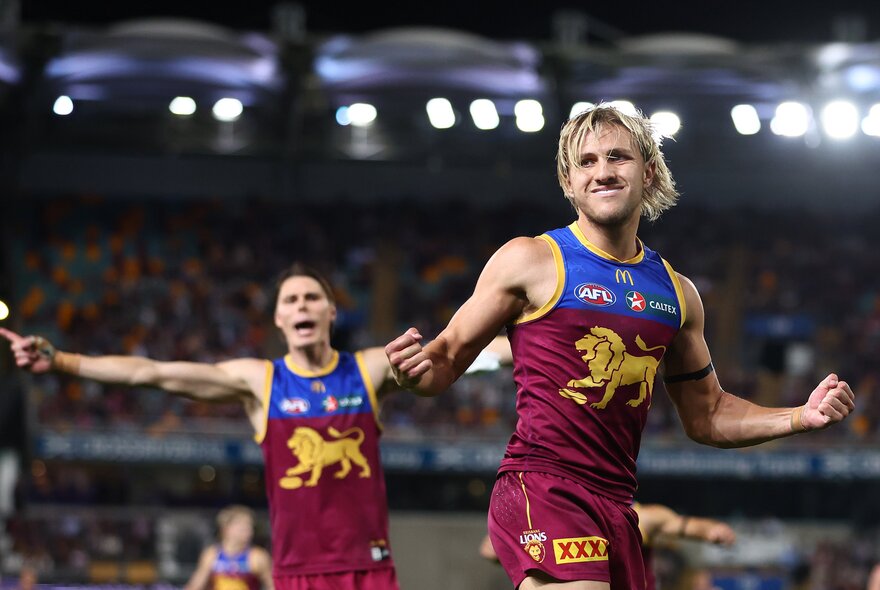 Two Brisbane Lions AFL players celebrating on the field, with blurred fans in the stadium in the background.