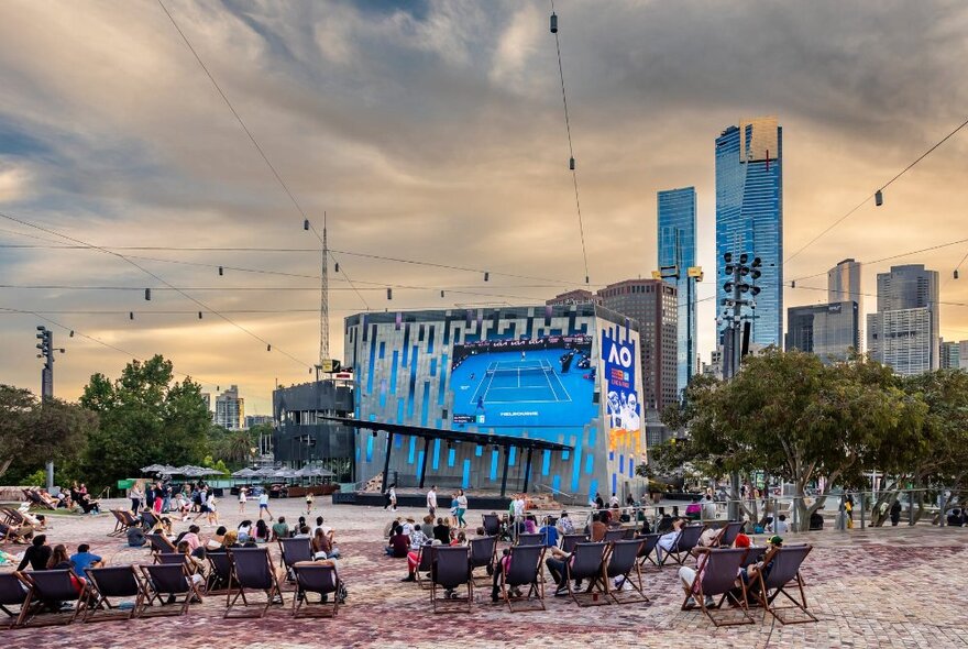 People seated in deck chairs at Federation Square watching the big screen with city buildings in the background.