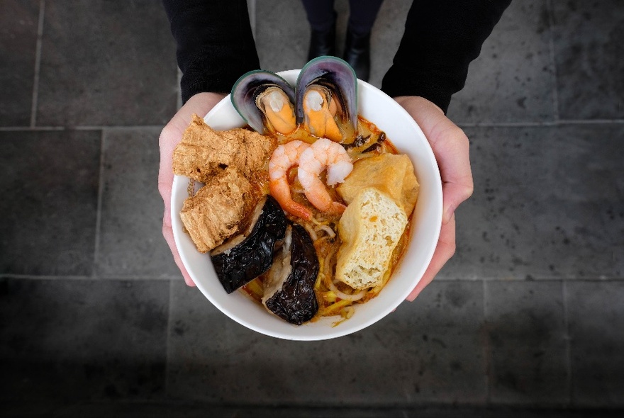 Bowl of laksa seen from above, hands and black-clad arms visible.