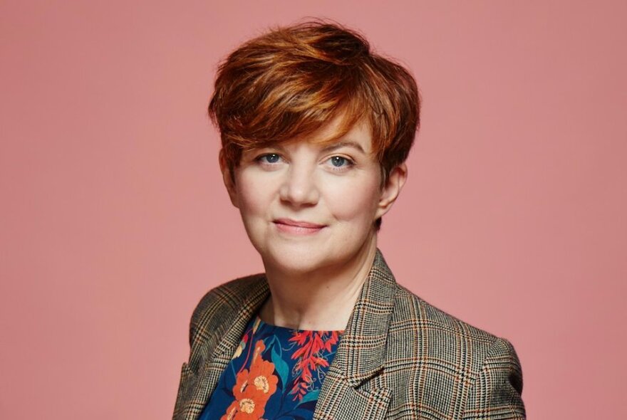 Filmmaker Sofya Gollan, a woman with short red hair, smiling slightly, wearing a floral top and a checked suit jacket, posed against a peach coloured backdrop.