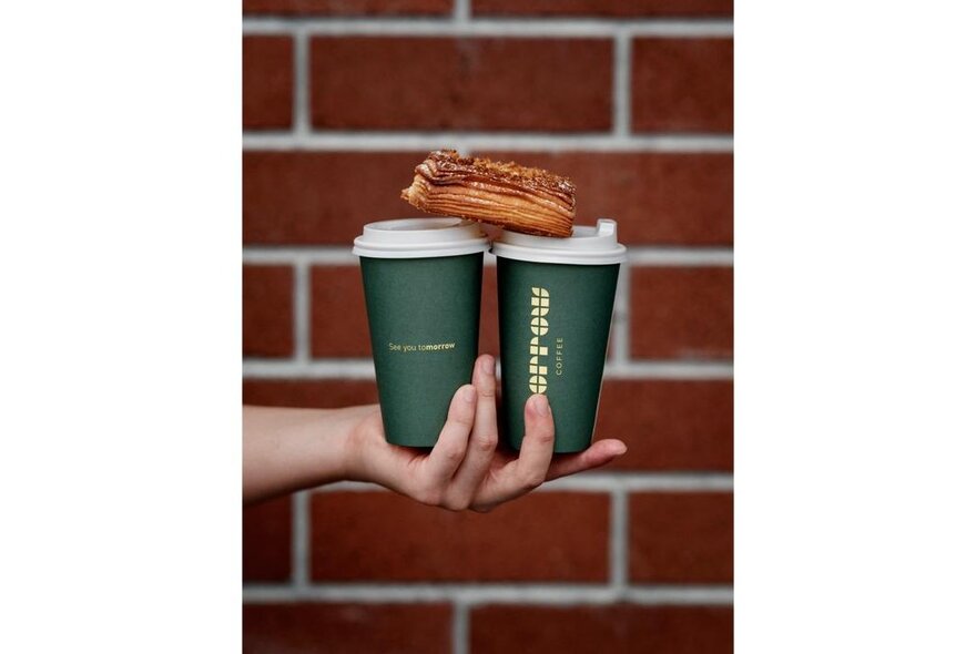 A person's hand holding two green takeaway coffee cups with a croissant balanced on top; red brick wall in the background.