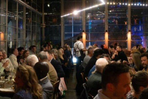 The dining room at Taxi Kitchen filled with people eating and drinking at tables, with large windows looking out onto the night sky.