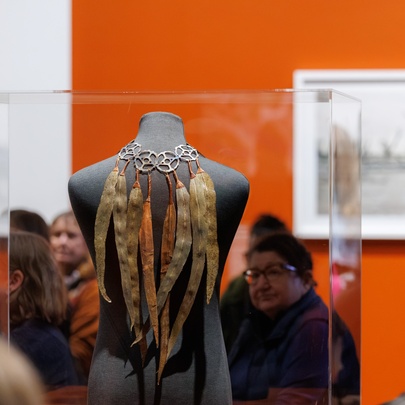 Museum display of a gum leaf necklace  in a glass case, people milling nearby in a room with one orange wall.