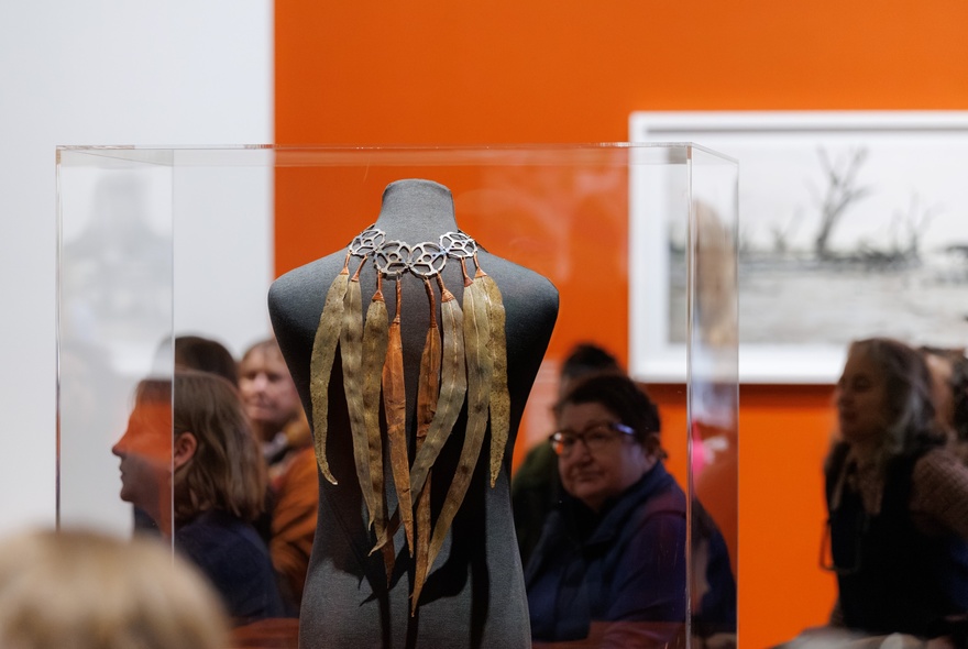 Museum display of a gum leaf necklace  in a glass case, people milling nearby in a room with one orange wall.