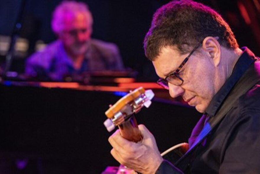 A man in side profile view, concentrating on playing an acoustic guitar, with an out of focus piano player in the background.