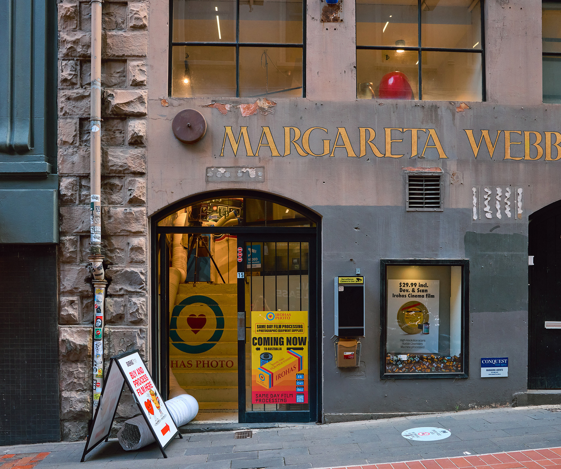 The exterior of a shop in an old building. There is a sandwich sign saying Irohas Photo Lab and a painted sign on the wall saying Margaret Webber.