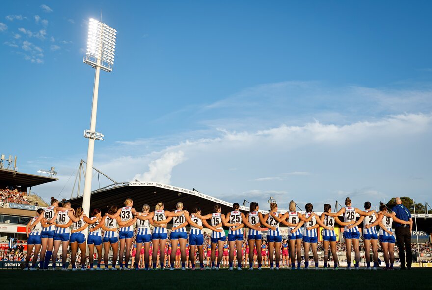 North Melbourne AFLW football team standing with arms linked on the Icon Park ground with light tower.
