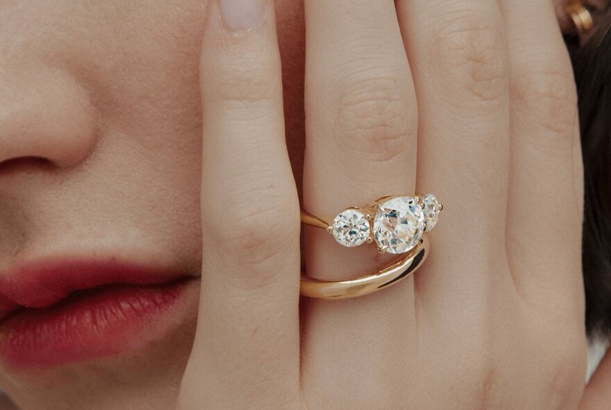 A close-up of a ring with three large diamonds and a gold hoop on a woman's hand as she holds it up to her face