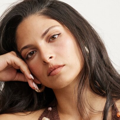 A close-up portrait of a young woman with long, dark brown hair resting her head gently on her right hand.