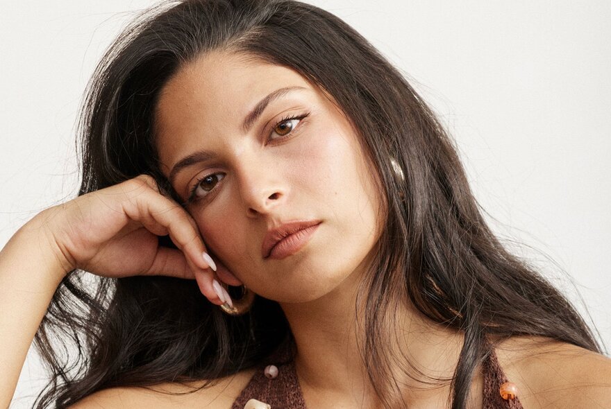 A close-up portrait of a young woman with long, dark brown hair resting her head gently on her right hand.
