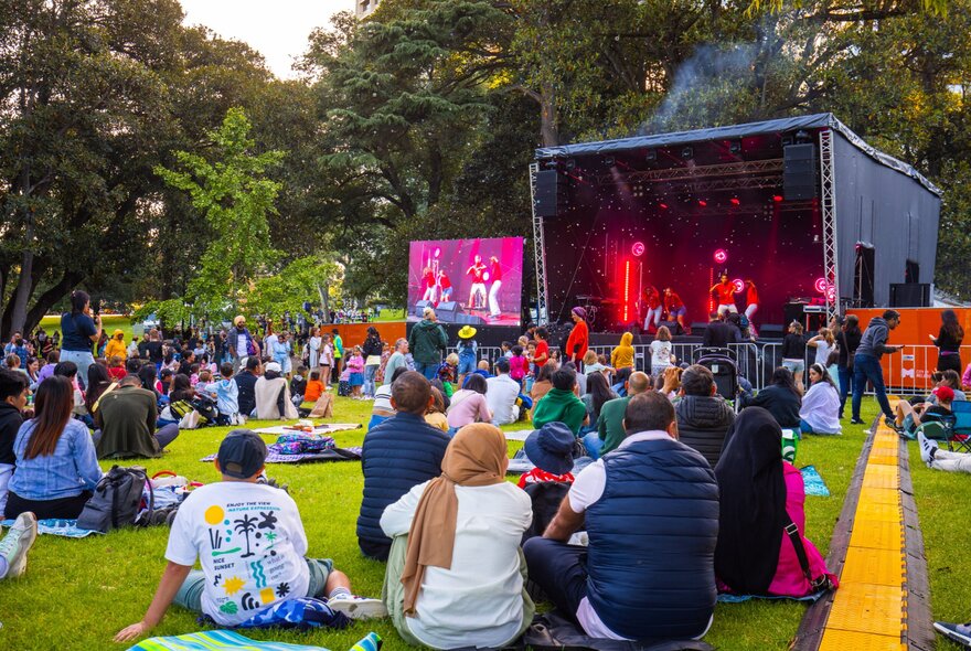 A crowd seated on the grass in front of a temporary stage in a park; trees in the distance.