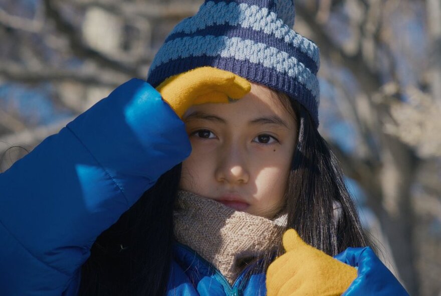 A child wearing winter clothing, shading her eyes from the sun with gloved hand.