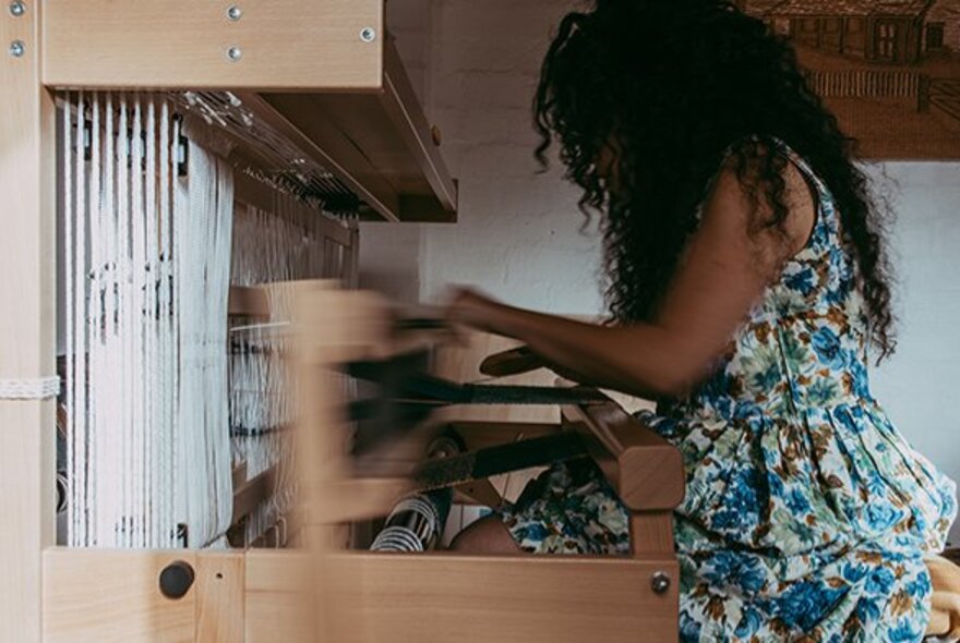 Woman weaving at a wooden hand loom, wearing a floral dress.