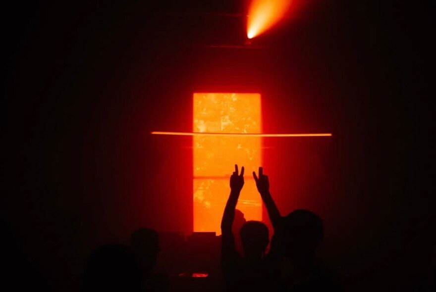 A DJ has their hands in the air in front of a rectangle of orange light in a very dark club. 
