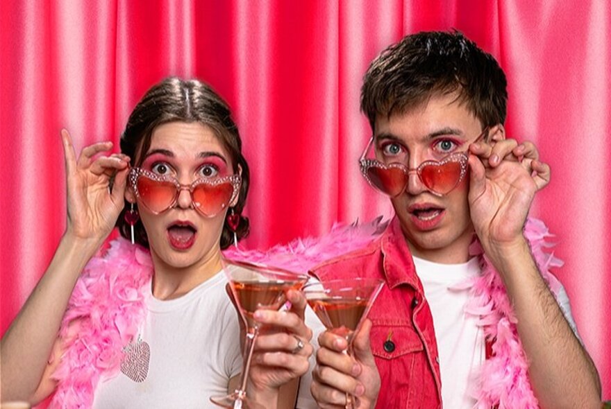 A young woman and man, both wearing pink heart-shaped sunglasses, pink feather boas and holding pink cocktails, in front of a pink curtain.