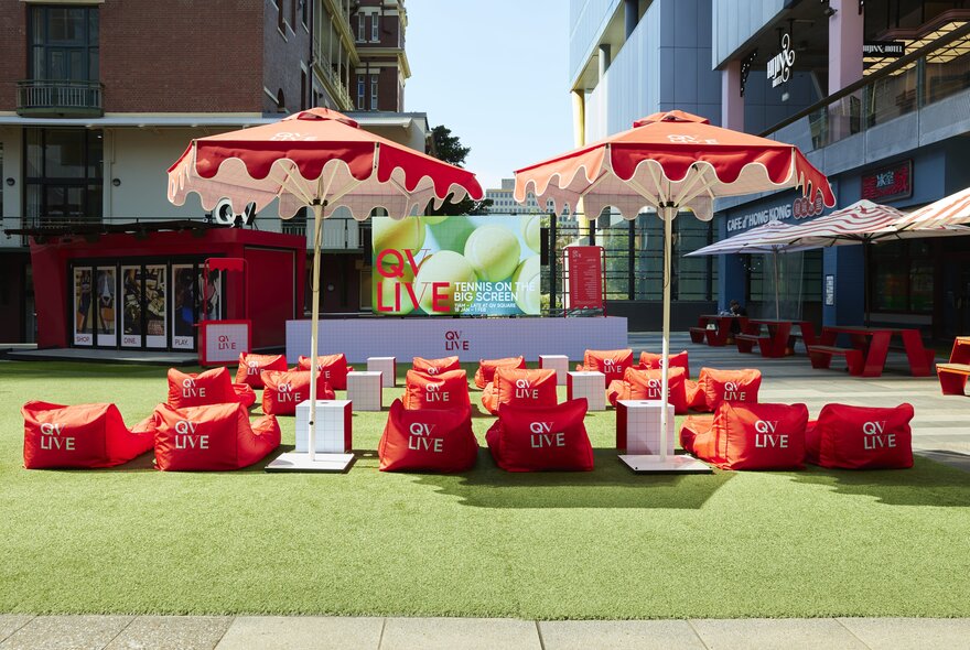 A large TV screen with red bean bag chairs under red fringed umbrellas on a surface of fake grass.