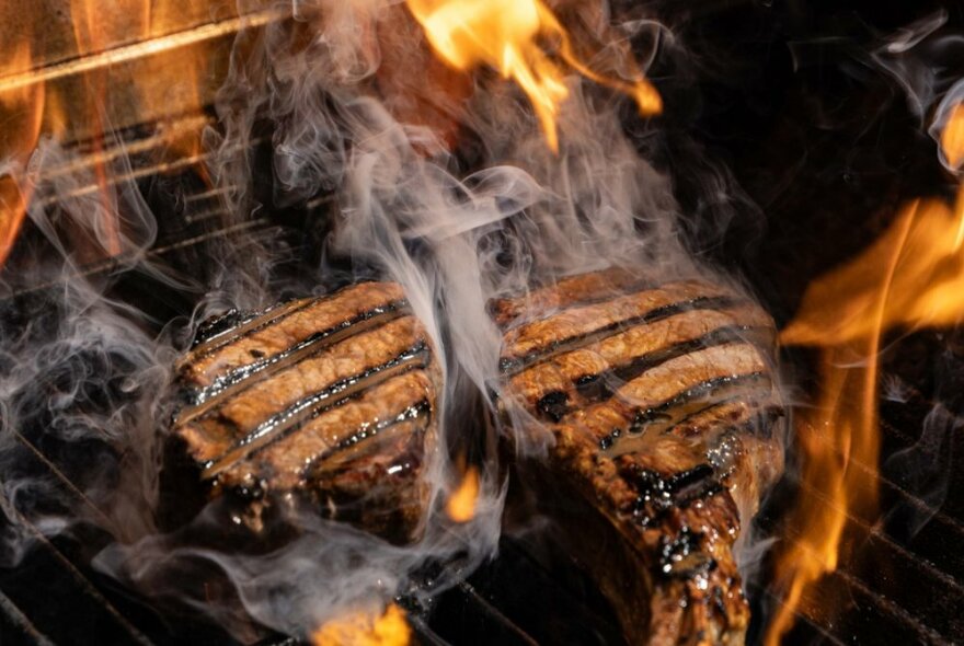 Two steaks on a char grill with grill marks visible, smoke and flames surrounding them. 