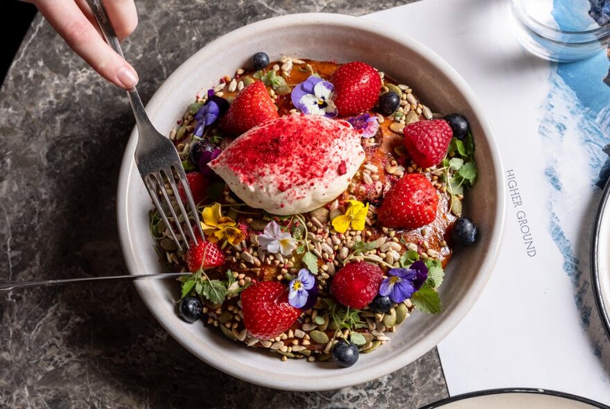 Colourful breakfast dish of granola with fuit and yoghurt scattered with flowers and pink powder.