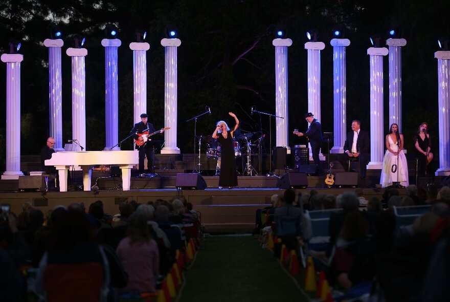 A woman on an outdoor stage with purple pillars and musicians behind her, a seated audience in front.