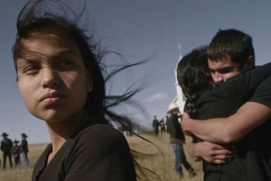 A still from the film Songs My Brothers Taught Me; on a large grassy plain a child is in the foreground, behind her a couple are hugging, with more people in small groups behind them.