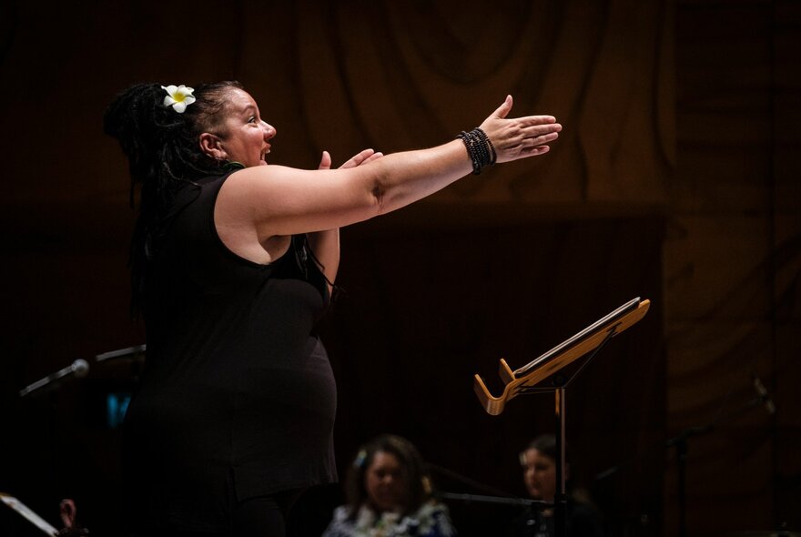 A woman in black performing with her arm straight out and a frangipani in her hair. 