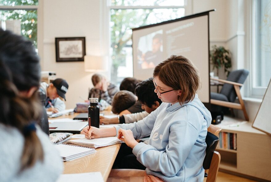 A bright and airy studio room used as a classroom, with children sitting and working around tables, windows and a large screen in the background.