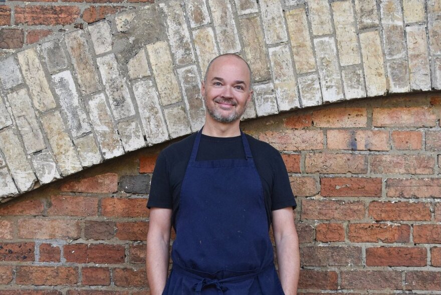 Chef Tobie Puttock wearing an apron and smiling, standing in front of a brick wall.