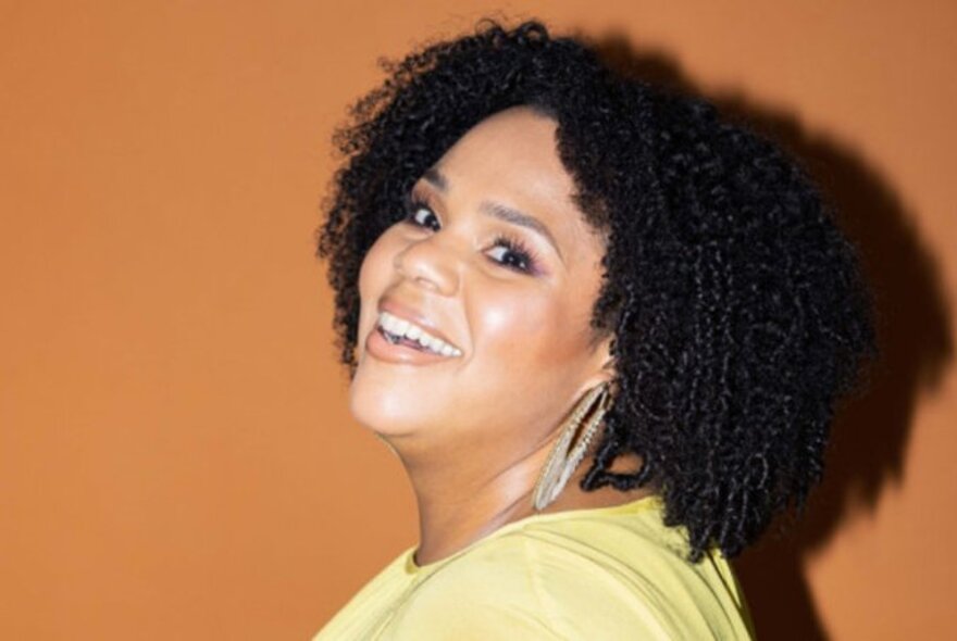 Comedian Desiree Burch, smiling with her mouth slightly open, a large earring dangling onto her shoulder, tightly curled black hair, wearing a pale yellow top, posing against a mustard coloured backdrop.