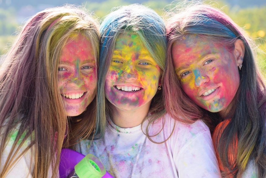 Three teenagers smiling at the camera, their faces covered in brightly coloured powder, at a Holi Festival.