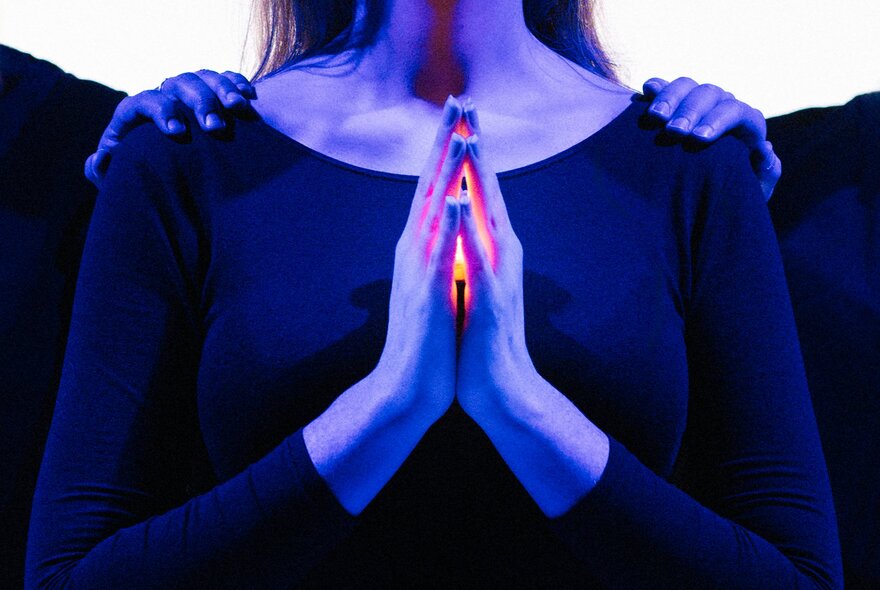 The torso of a woman wearing a black top, hands pressed together in prayer with a bright orange glow coming from between them; two hands resting on her shoulders.