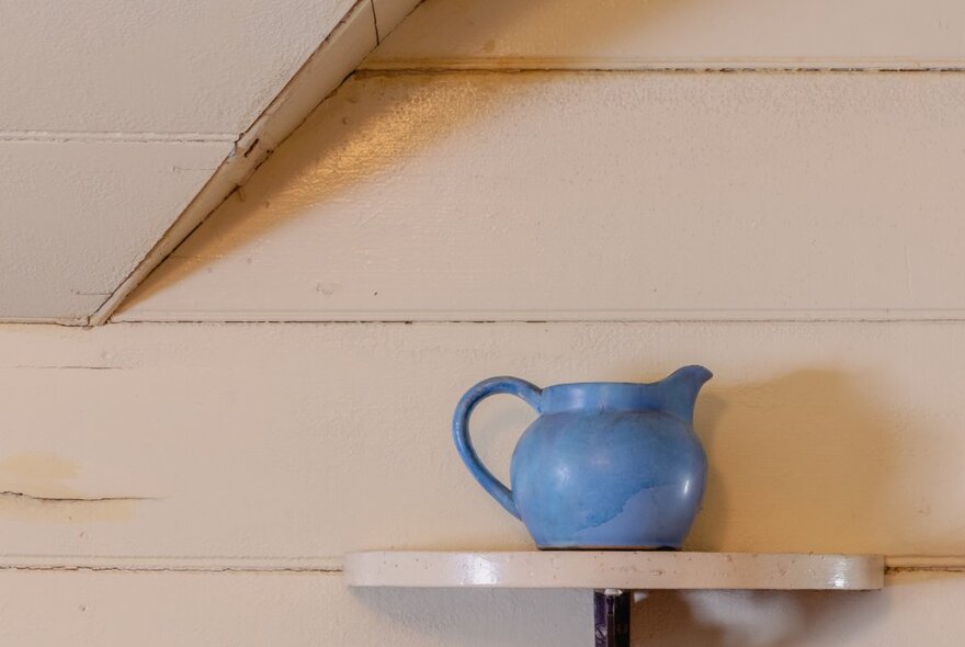 A squat blue ceramic jug on a small half-circle table against a cream-painted panelled wall.