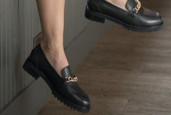 Feet wearing black flat casual shoes with a gold front chain link detail, with bare legs.
