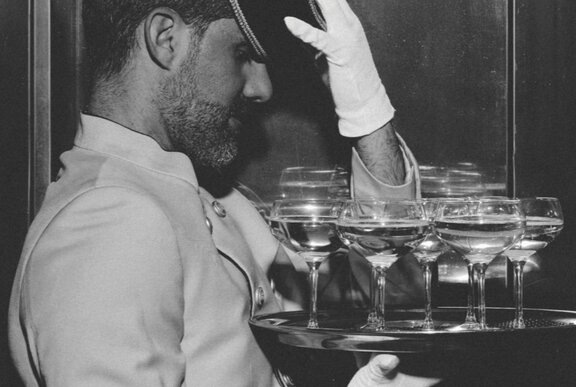 Black and white photograph of a formally-dressed waiter with white gloves holding a tray of wide-mouthed champagne glasses.