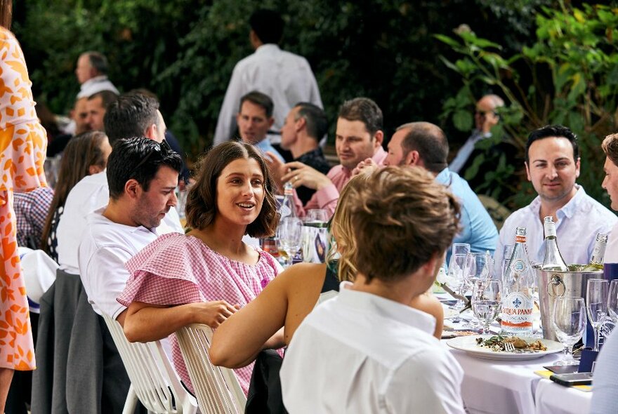 People seated, talking at a long table laden with food and drinks, outdoors.